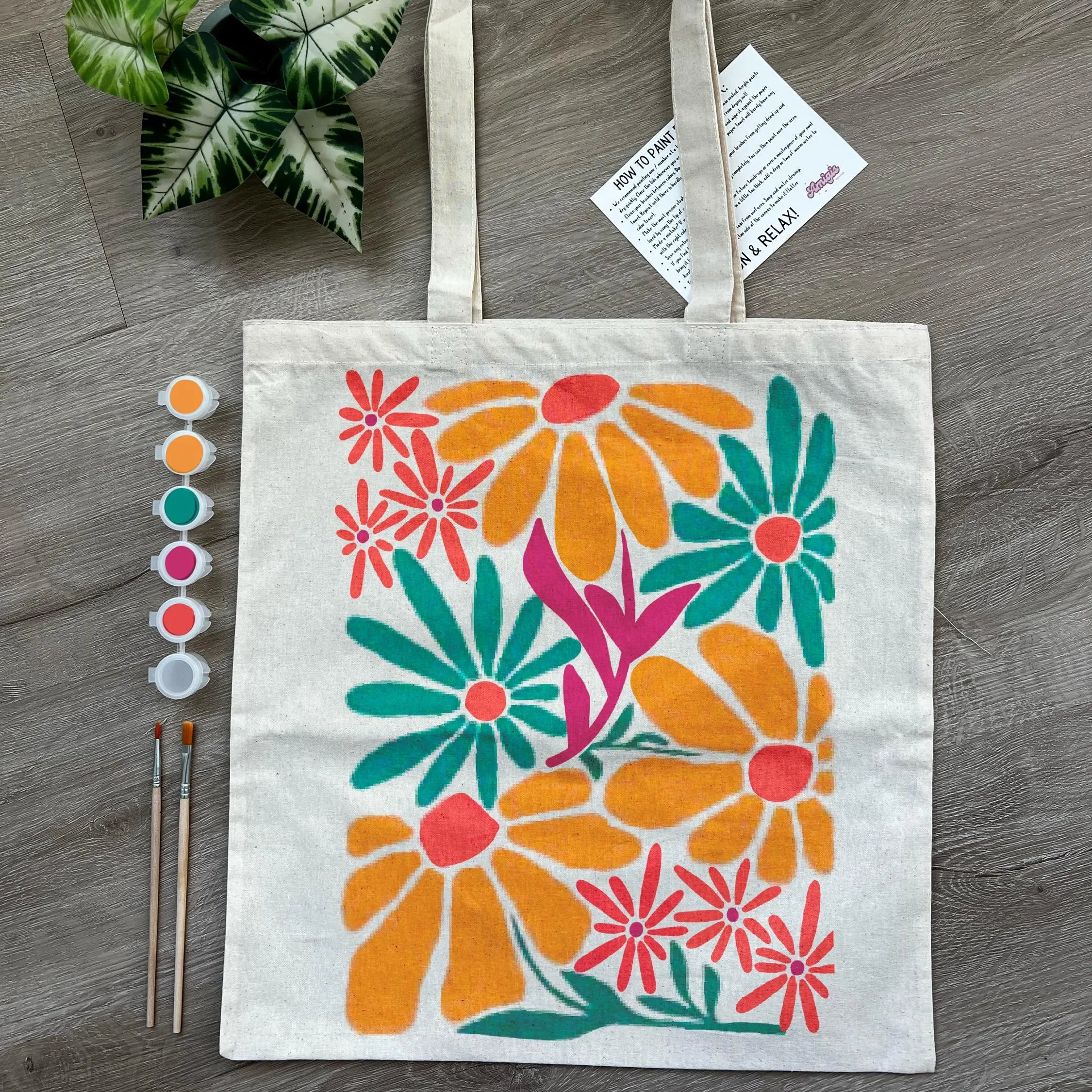 Canvas tote bag with colorful floral design on a wooden surface, accompanied by paint samples and brushes.